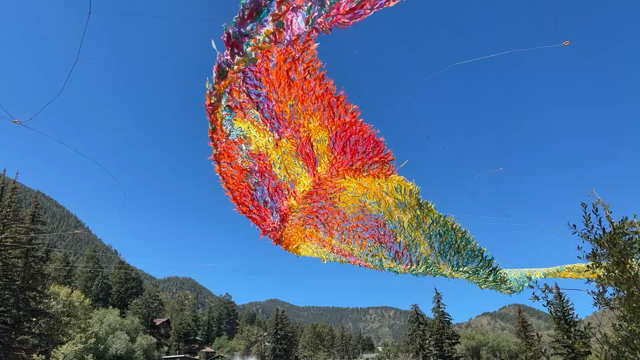 A fluttering, undulating sheet like form, made from strips of kite material in a color gradient going from warm to cool colors. In the background is a blue sky and a mountainside filled with evergreen trees.