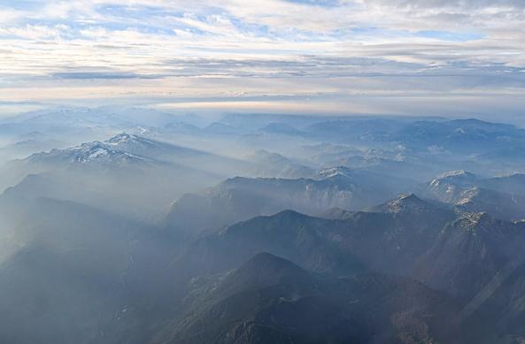 From an airplane, slanting sunbeams over big mountains. A bit of blue above, among the clouds.