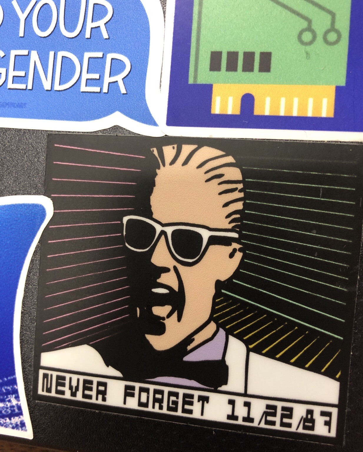 Back of a laptop with stickers. Primarily in view is wbm312’s Max Headroom “11/22/87 Never Forget” sticker.