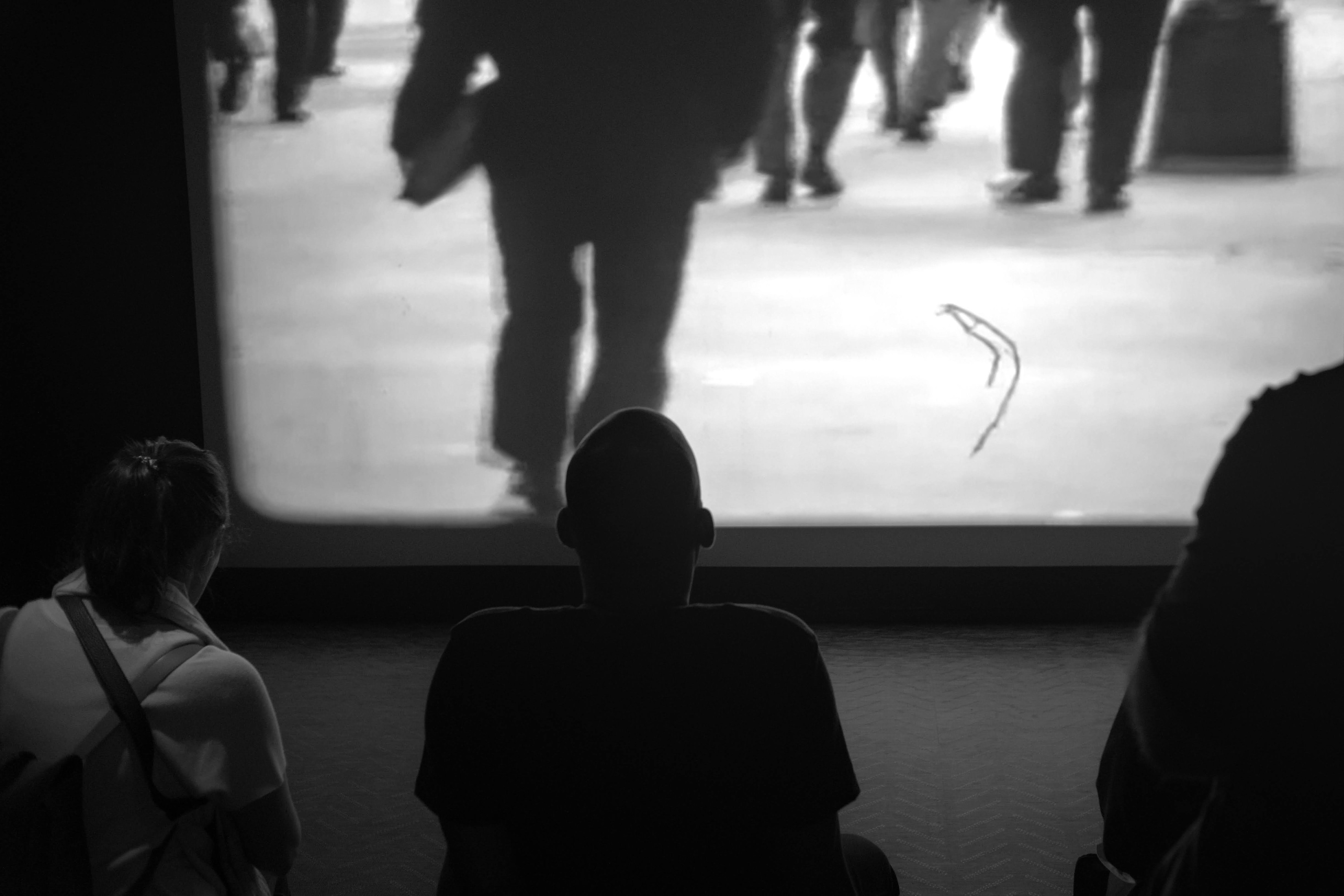 A black and white photo of museum goers looking up at a projected film created from Maier’s recordings. 