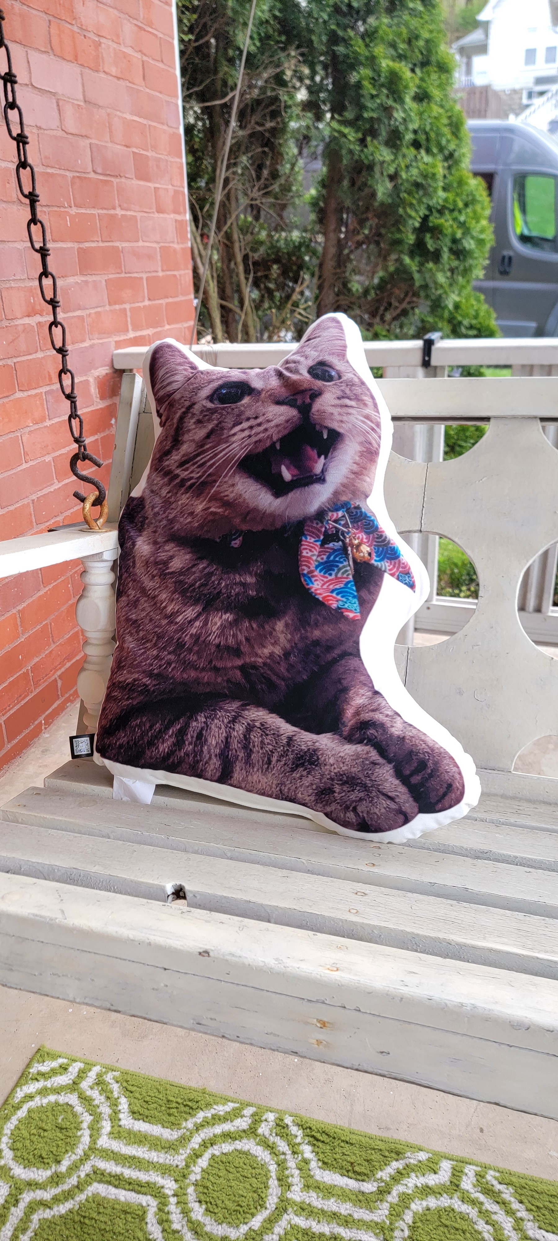 A very large pillow of a tabby cat is sitting upright on a white porch swing in front of a red brick house. The cat looks humorously large.