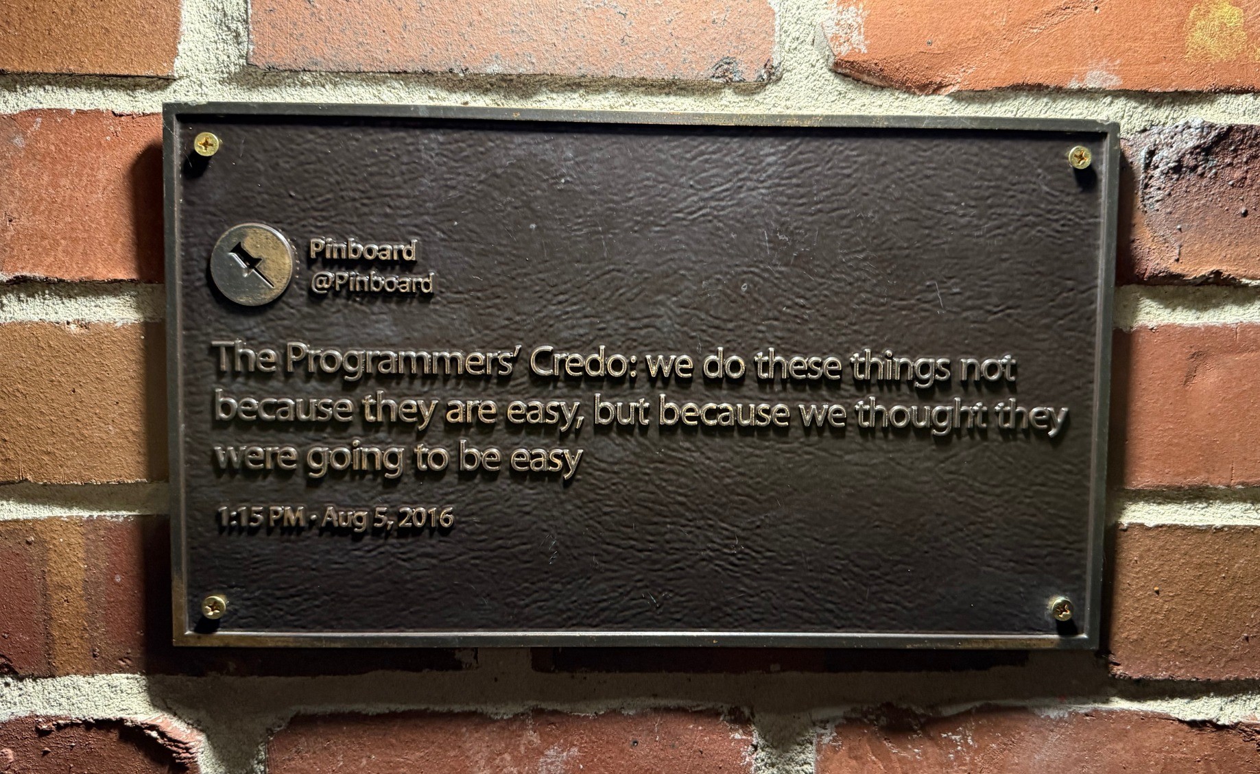A museum quality brass plaque replica of a Twitter post from August 5th, 2016 -- "The Programmers' Credo: we do these things not because they are easy, but because we thought they were going to be easy". It is mounted via brass screws on a brick wall. The picture was taken at night, lighted from directly above.