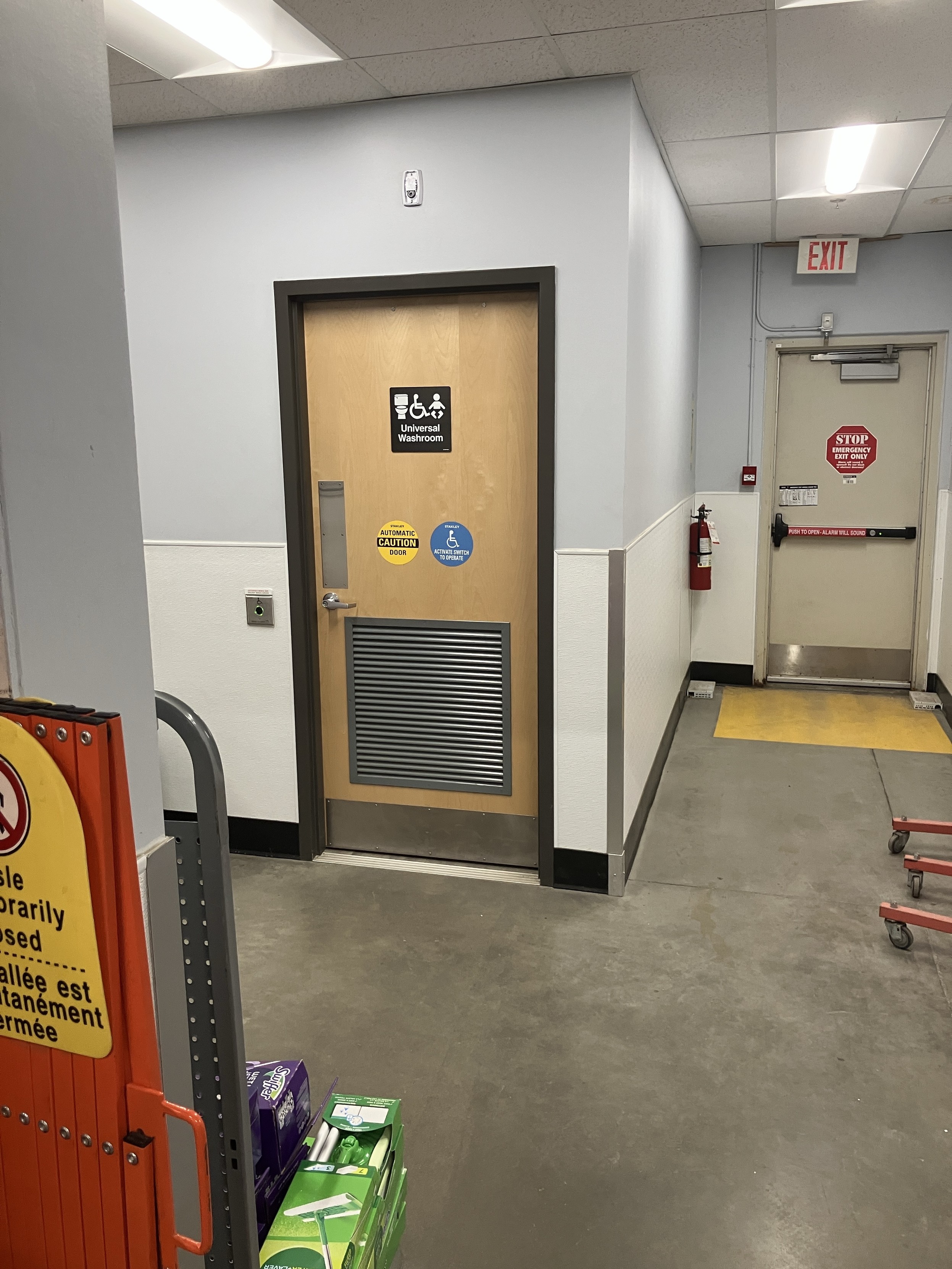 A hallway featuring a door labeled "Universal Washroom," with accessibility symbols and caution signs. Nearby, there is a fire extinguisher, an "Exit" sign, and a sign indicating a temporarily closed aisle.