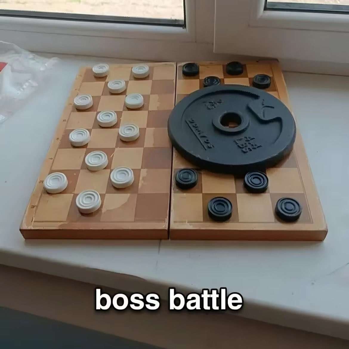 Photo of a game of checkers that has been setup next to a windows, except there is a 2 kg (4.4 lbs) weight disc covering a 4x4 square area on the side with the black pieces. Caption at the bottom says:
boss battle