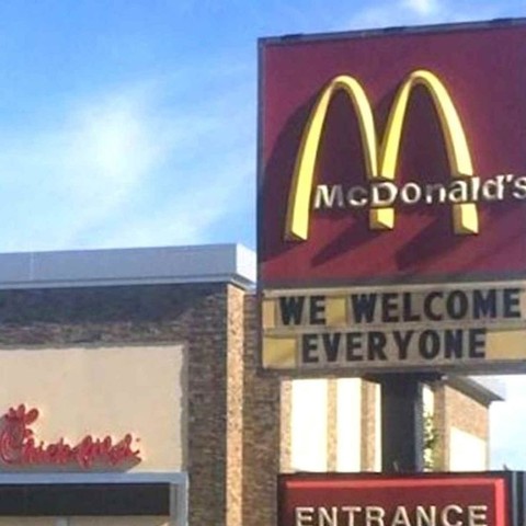 The image captures a daytime exterior view of a McDonald's restaurant, along with a portion of what appears to be a neighboring Chick-fil-A. The focus is primarily on the McDonald's signage, which dominates the right side of the frame.

The McDonald's sign is a classic design featuring the iconic golden arches in a bright yellow hue against a maroon background. Below the arches, the word "McDonald's" is printed in a silver font. Beneath the primary logo, a marquee board displays the message "WE…
