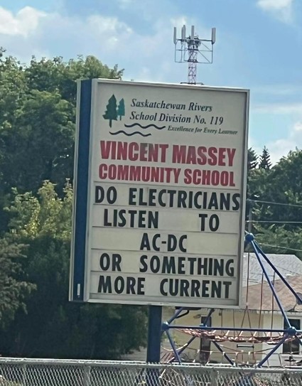 The image shows a sign for Vincent Massey Community School in front of a backdrop of trees and a cloudy sky. The sign is rectangular with a light background and dark blue borders. At the top of the sign, there is the logo for Saskatchewan Rivers School Division No. 119, including a stylized image of trees and waves. The words "Excellence for Every Learner" are written below the logo. The main text of the sign reads "VINCENT MASSEY COMMUNITY SCHOOL" in large, red lettering. Below this is a quest…