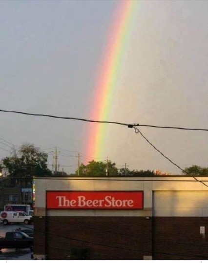 The image captures a striking scene featuring a rainbow arching over a "The Beer Store" establishment. The sky is predominantly a pale gray, hinting at overcast weather. A vibrant rainbow dominates the upper portion of the frame, displaying a full spectrum of colors from red to violet. The bottom of the rainbow appears to touch down just beyond the roof of the beer store, creating a humorous visual juxtaposition.

The beer store itself is a modern, rectangular building with a red rectangular si…