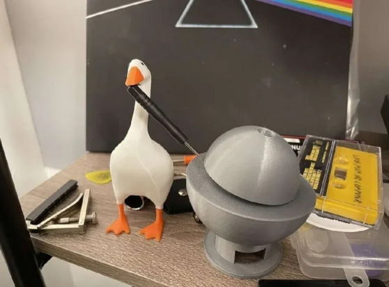 The image showcases a whimsical setup on a wooden surface, likely a desk or shelf. The central focus is on a 3D-printed goose figurine, its white body and orange beak and feet standing out against the other elements. The goose is holding a black screwdriver in its beak, appearing to "work" on a grey, 3D-printed spherical object. This sphere is composed of two parts, a top and a bottom section, and has a small hole on the top.

Other items are scattered around the scene. To the left of the goose…