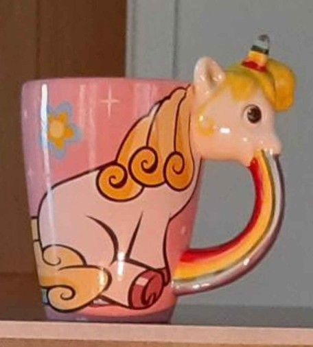 Photo of a coffee mug with a unicorn on it. The unicorn head throwing up a rainbow make up the handle.