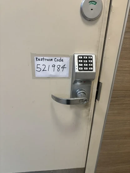 The image showcases a portion of a restroom door. The door is painted in a light, off-white color and features a combination lock and a handle.

A handwritten sign, taped to the door, indicates "Restroom Code" followed by the number "521984". The sign is made of plain white paper and the text is written in black marker. The combination lock is a metallic gray color and has a keypad with numbers 1 through 9, an asterisk, and a zero. Above the lock is a circular indicator with the word "VACANT" v…