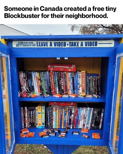 The image shows a blue, small, public video library box with glass doors, situated outdoors. The box is labeled "LEAVE A VIDEO • TAKE A VIDEO" at the top, with a sign below that reads "BE KIND PLEASE REWIND MAKE IT A BLOCKBUSTER NIGHT." Inside, there are two shelves filled with a variety of DVDs and VHS tapes, including titles like "The Hunger Games," "The
 Goonies," and "The Matrix." The bottom shelf also contains an assortment of candy bars, including Reese's and Snickers, alongside batteries…
