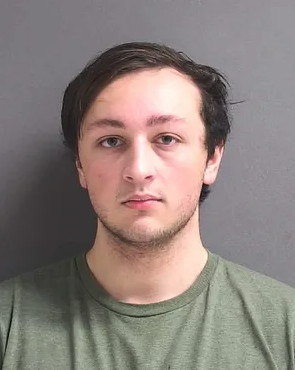 A mugshot of then 19-year-old Noah Michael Urban. He has brown hair, combed over the right side of his face. He is somewhat unshaven and wearing a green t-shirt.