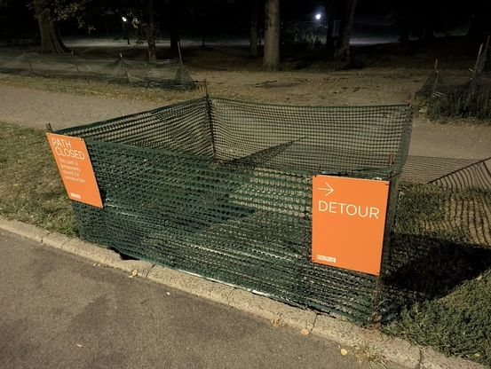 A photo at night of a fenced off segment of grass, roughly 2m x 2m, between a road and a path that parallels it. There is no path enclosed in the fence, which has two signs.

On the left, one sign reads "PATH CLOSED. This path is temporarily closed for construction." The sign on the right simply says "DETOUR" and has an arrow pointing to the right, even though the sign is at the right edge of the fenced area.