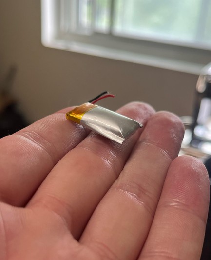 Photo of someone holding a tiny spicy pillow (lithium ion battery that has swollen and will explode soon). The battery from a USB device, which is small enough to be held with one fingers, has a red and black wire on one side with gold colored tape and a tiny bit of white smoke coming out. Well that's not good...