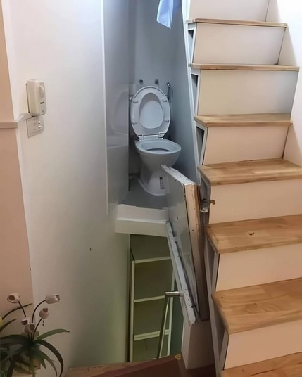 Photo of a toilet that's been squeezed by some stairs on the middle step area, but across a gap. To get over there, there's a trap door style walkway above some stairs that go to the floor below. Hope that thing never overflows or leaks.