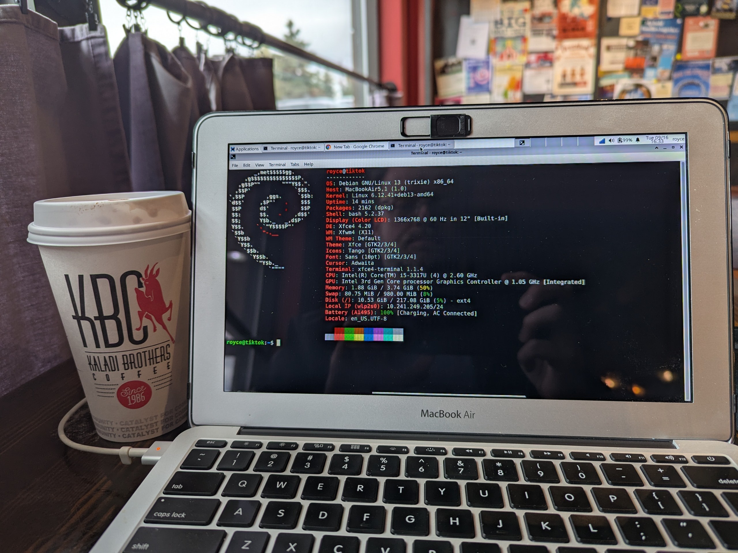 Macbook sitting on table at coffee shop, showing fastfetch output: Debian 13 Trixie, MacBook Air 5,1, 4G RAM, resolution 1366 x 768, CPU Intel Core i5-3317U, 220GB SSD. Kaladi Brothers coffee cup to the left with red jumping goat logo. Community bulletin board and window in the background. Bash prompt shows Royce at tiktok (the robot from The wizard of Oz, not the social media app)