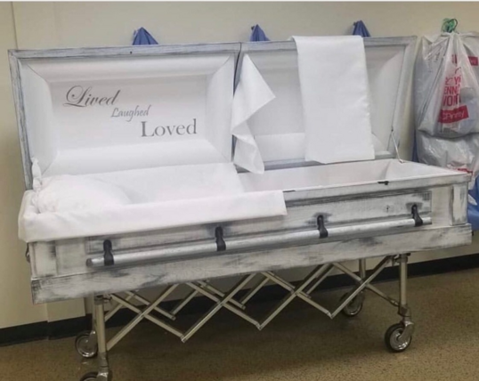 The image showcases an open casket, likely in a funeral home or similar setting. The casket is the central focus, positioned on a wheeled stand. The casket's exterior has a distressed, weathered appearance with a predominantly white base and a hint of gray, giving it a rustic or vintage aesthetic. The sides of the casket feature black handles.

The interior of the casket is lined with pristine white fabric. The head area of the casket is adorned with the words "Lived, Laughed, Loved" in a delic…