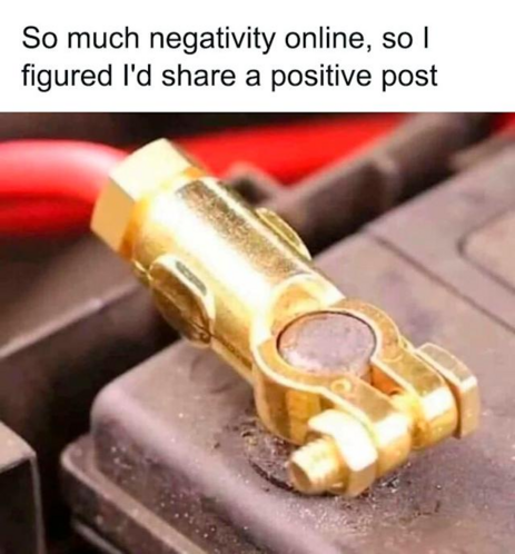 The image is a close-up photograph of a gold-colored car battery terminal connector. The connector is the central focus, positioned slightly off-center. It appears to be made of brass or a similar metal, with a shiny, polished finish. The connector has a cylindrical body with a hexagonal nut at the top and a circular eyelet on the bottom. A smaller bolt is visible on the side of the connector, used to secure it to the battery terminal.

The connector rests on a dark gray, textured surface that …