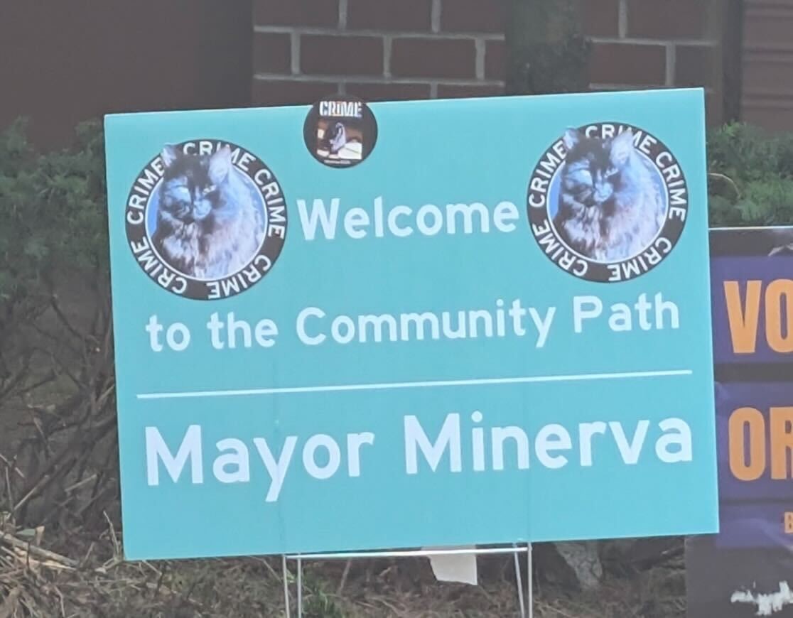 A teal sign with centered white lettering: Welcome
to the community path
Mayor Minerva
On either side of “welcome” are pics of a black longhaired cat with lettering around the pics that say “CRIME” over and over.