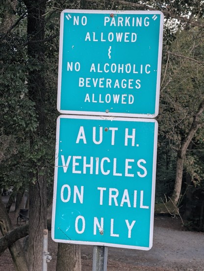 The image shows two green road signs with white text, mounted on a metal pole. The top sign reads: "NO PARKING ALLOWED & NO ALCOHOLIC BEVERAGES ALLOWED." The bottom sign states: "AUTH. VEHICLES ON TRAIL ONLY." The signs are positioned in a natural setting with trees and a dirt path visible in the background. The signs appear to be weathered, with some visible wear and tear, including chipped paint and minor scratches. The text is clear and legible, with the top sign having quotation marks aroun…