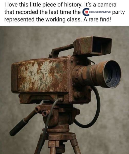 The image features a close-up of an antique, heavily rusted movie camera mounted on a tripod. The camera is the focal point, taking up the majority of the frame. It has a boxy body with a viewfinder on top and a large lens at the front. The camera's surface is covered in rust, with areas of peeling paint and discoloration, suggesting significant age and exposure to the elements. The tripod is also rusted and appears to be made of the same metal as the camera, with three legs and a mechanism for…