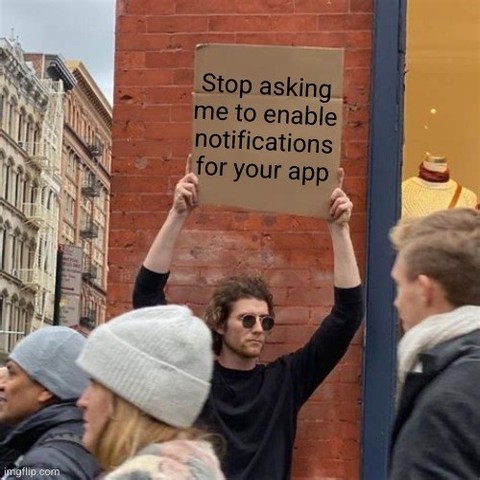 The image depicts a man holding a cardboard sign in an urban setting. The sign reads "Stop asking me to enable notifications for your app." The man is the focal point of the image, positioned centrally and facing the viewer, though his gaze is directed slightly to the right. He has light skin, curly brown hair, and is wearing sunglasses. He is dressed in a black long-sleeved shirt. His arms are raised, holding the sign above his head.

The background suggests a city street scene. To the left of…