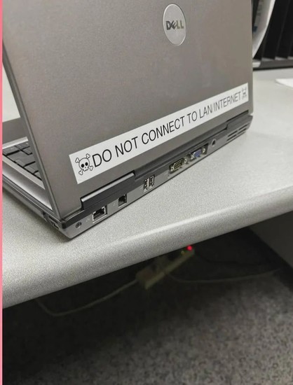 The image features a gray Dell laptop sitting on a light gray desk. The laptop is slightly open, revealing a portion of the keyboard. The most prominent feature is a white sticker affixed to the top of the laptop's lid, near the hinge. The sticker has black text that reads "DO NOT CONNECT TO LAN/INTERNET" and features a small skull and crossbones graphic at the beginning of the sentence.

The laptop's back edge, where the ports are located, is visible. There are various ports and connectors, in…