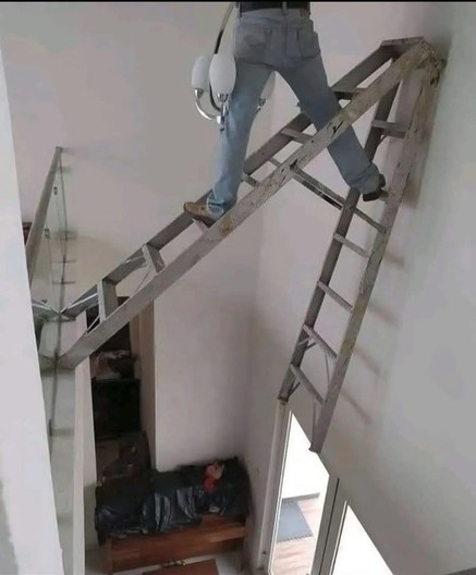 Unhinged photo of a "handyman" replacing a light fixture by just propping a aluminum ladder across a second story gap. The top of the ladder is just resting on the drywall while one part of the bottom is being prepped up on a small ledge with a glass railing.
