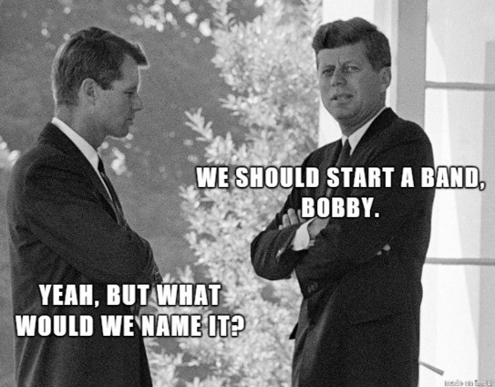 The image is a black and white meme featuring two men, likely John F. Kennedy and Robert Kennedy, in a humorous conversation. The scene appears to be outdoors, with a background of blurred foliage and what seems to be a building facade on the right.

The man on the left is seen from the side, wearing a dark suit and tie. His arms are crossed, and his expression is thoughtful.

The man on the right is facing the viewer, also in a suit and tie. He has his arms crossed and is looking towards the v…