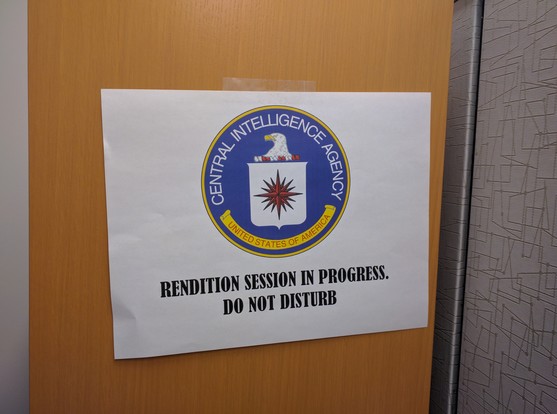 The image shows a rectangular white sign taped to a wooden door. The sign features the official seal of the Central Intelligence Agency (CIA) at the top. The seal is circular, with a blue outer ring containing the words "CENTRAL INTELLIGENCE AGENCY" in white lettering. Inside the ring is a yellow band with "UNITED STATES OF AMERICA" written in black. The central emblem within the seal depicts a white eagle with a red and white shield, and a red starburst at the center. Below the seal, in bold b…