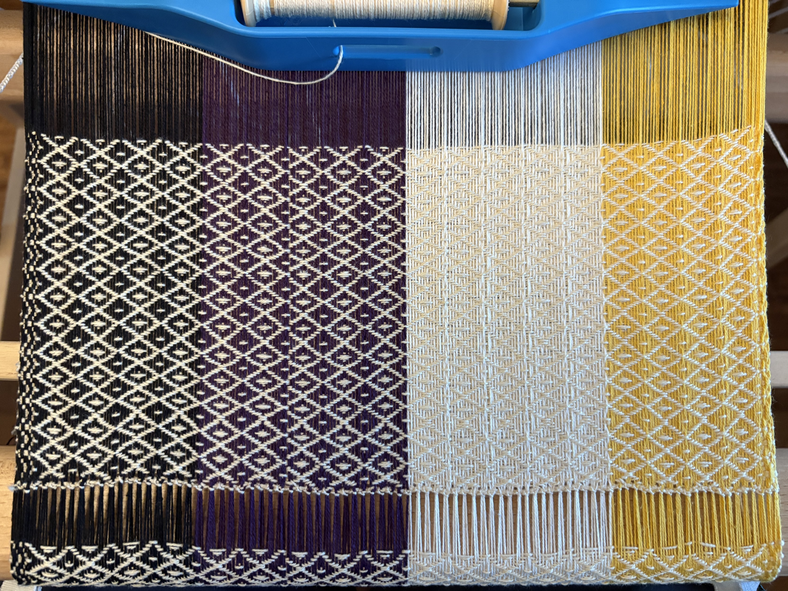 Weaving with a warp of black, purple, white, and yellow stripes (as in nonbinary pride flag) with white yarn as the weft making a diamond pattern