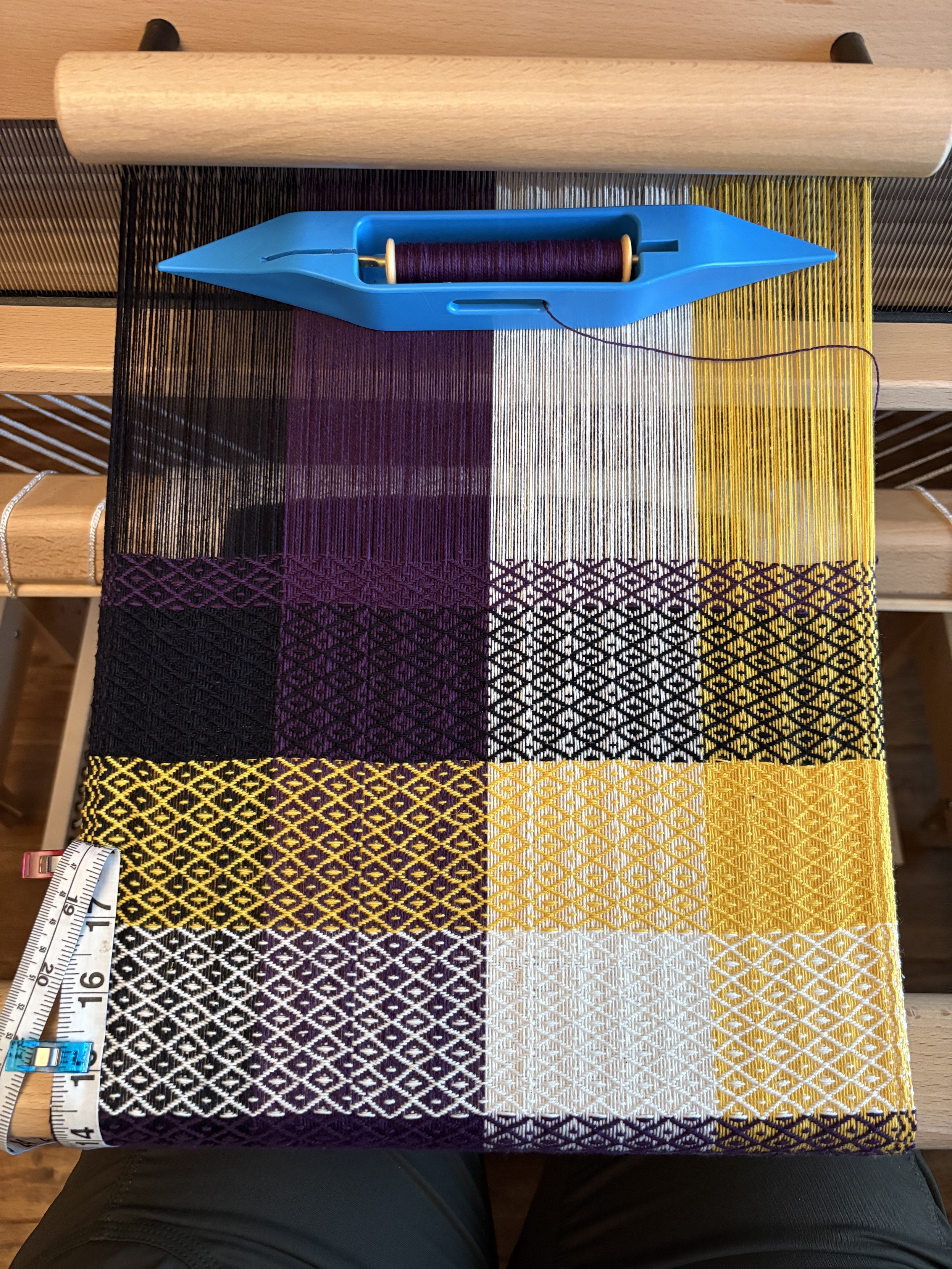 A towel in progress on a loom. The warp is black, purple, white, and yellow stripes. The weft is stripes of the same color, creating something of a checkerboard effect, with a diamond pattern.