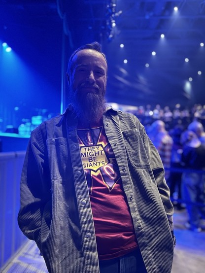 The image shows a person standing in front of a blurred, dark background, seemingly a crowd at a concert or event. The person is centered in the frame and facing the camera. They have short, dark hair and a thick, dark beard. The person is wearing a dark, long-sleeved shirt with a lighter colored graphic on the front, layered under a light-colored denim jacket. The background is out of focus, displaying indistinct shapes and colors suggesting a large group of people. The person’s expression is …