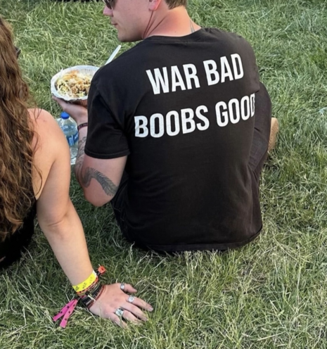 Man sitting with a woman facing away from the camera. 

On the back of the man's shirt are the words 

WAR BAD
BOOBS GOOD