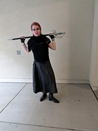 White woman with red hair and dark black eye makeup is wearing black clothes with bare arms and fingerless gloved hands resting on a rapier over her shoulders