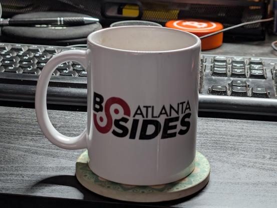 A white ceramic mug sits on a green coaster in front of a blurred background of a computer keyboard and other objects. The mug is cylindrical with a handle on the right side. A bold, red design is printed on the front of the mug, consisting of the words "BSIDES" stacked vertically. The text "BSIDES" is written in a stylized font with the "B" being significantly larger than the rest of the letters. The coaster is round and appears to be made of a textured material, possibly cork or a similar sub…