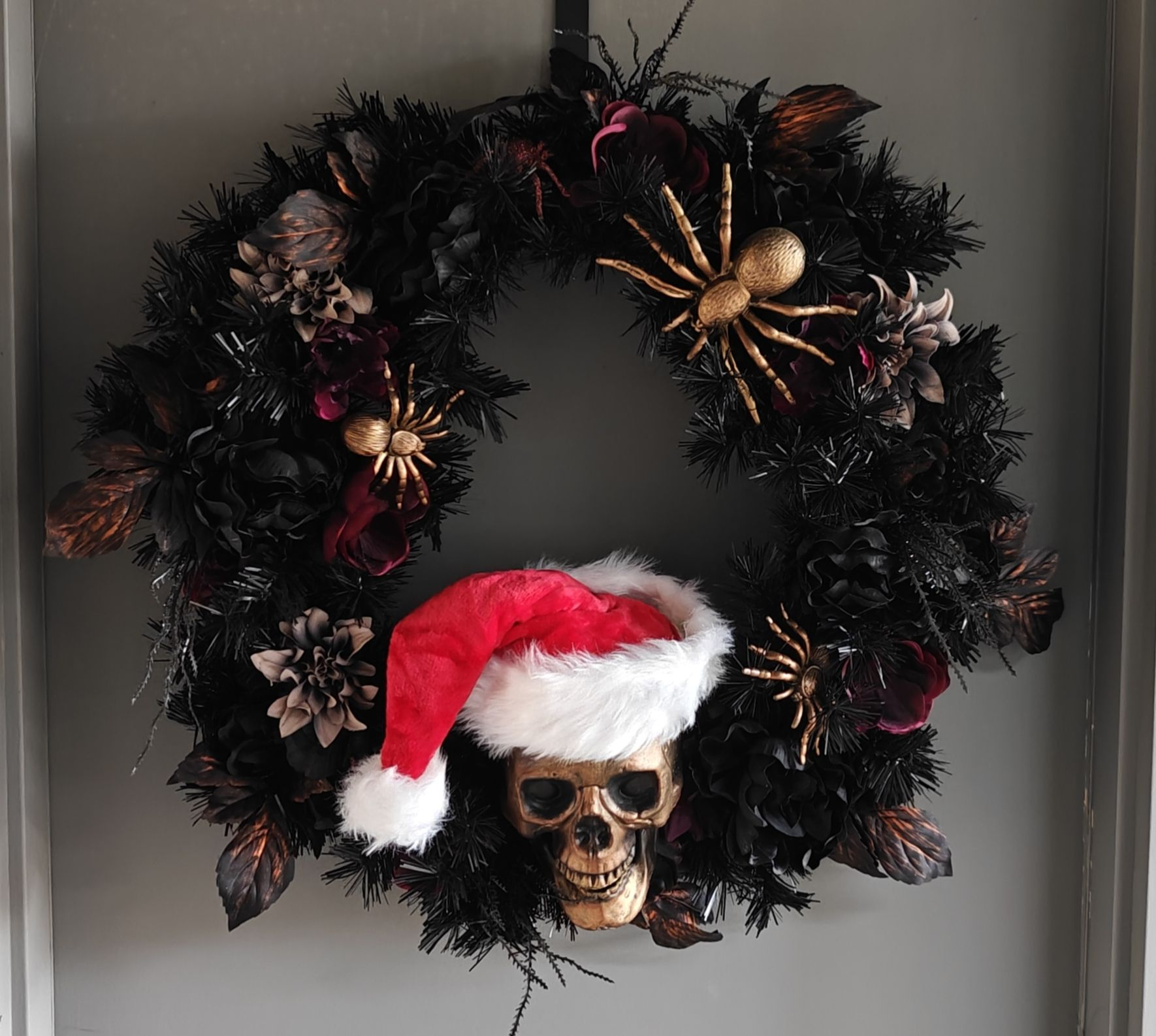 A Halloween wreath on an exterior door. It's all dark colors with some large spiders and a skull. The skull is wearing a Santa hat.