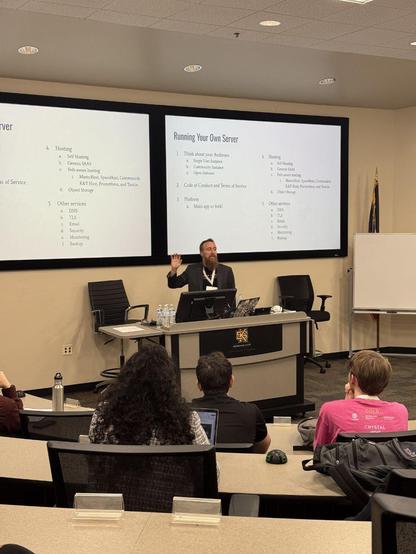 he image shows a conference room with a person standing at a podium giving a presentation. Two large screens display identical slides titled “Getting Started on the Fediverse”. In the foreground, a table is set with microphones and water bottles, and several people are seated facing the presenter. An American flag is partially visible in the left corner of the frame. 

The slides list the following points:

    “Pick a username”
    “Fill out your profile”
    “Set privacy preferences and enabl…