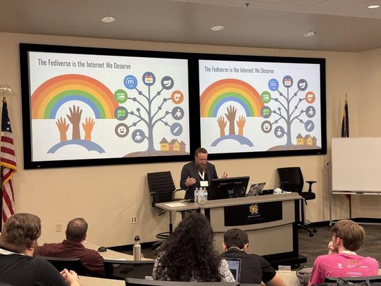 The image shows a person standing at a podium giving a presentation in a lecture hall. Three large screens behind the podium display presentation slides with bullet-point lists. Several people are seated in rows of chairs facing the podium, appearing to be listening to the presentation. 
