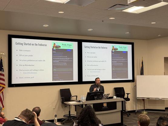 The image shows a person standing at a podium giving a presentation in a lecture hall. Three large screens behind the podium display presentation slides with bullet-point lists. Several people are seated in rows of chairs facing the podium, appearing to be listening to the presentation. 

The left screen reads "4. Hosting" with sub-points "Self Hosting", "Cloud Hosting", "Dedicated Hosting" and "Bare Metal Hosting". The center screen reads "Running Your Own Server" with sub-points "1. Think abo…