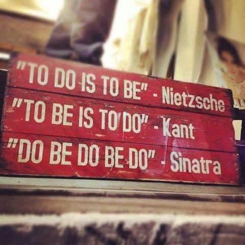 The image shows three rectangular red signs stacked on top of one another. The signs are angled slightly upward and appear to be made of metal. Each sign has white lettering. The top sign reads “TO DO IS TO BE” - Nietzsche. The middle sign reads “TO BE IS TO DO” - Kant. The bottom sign reads “DO BE DO BE DO” - Sinatra. The background is out of focus and reddish in tone with some indiscernible shapes suggesting furniture or a room interior.