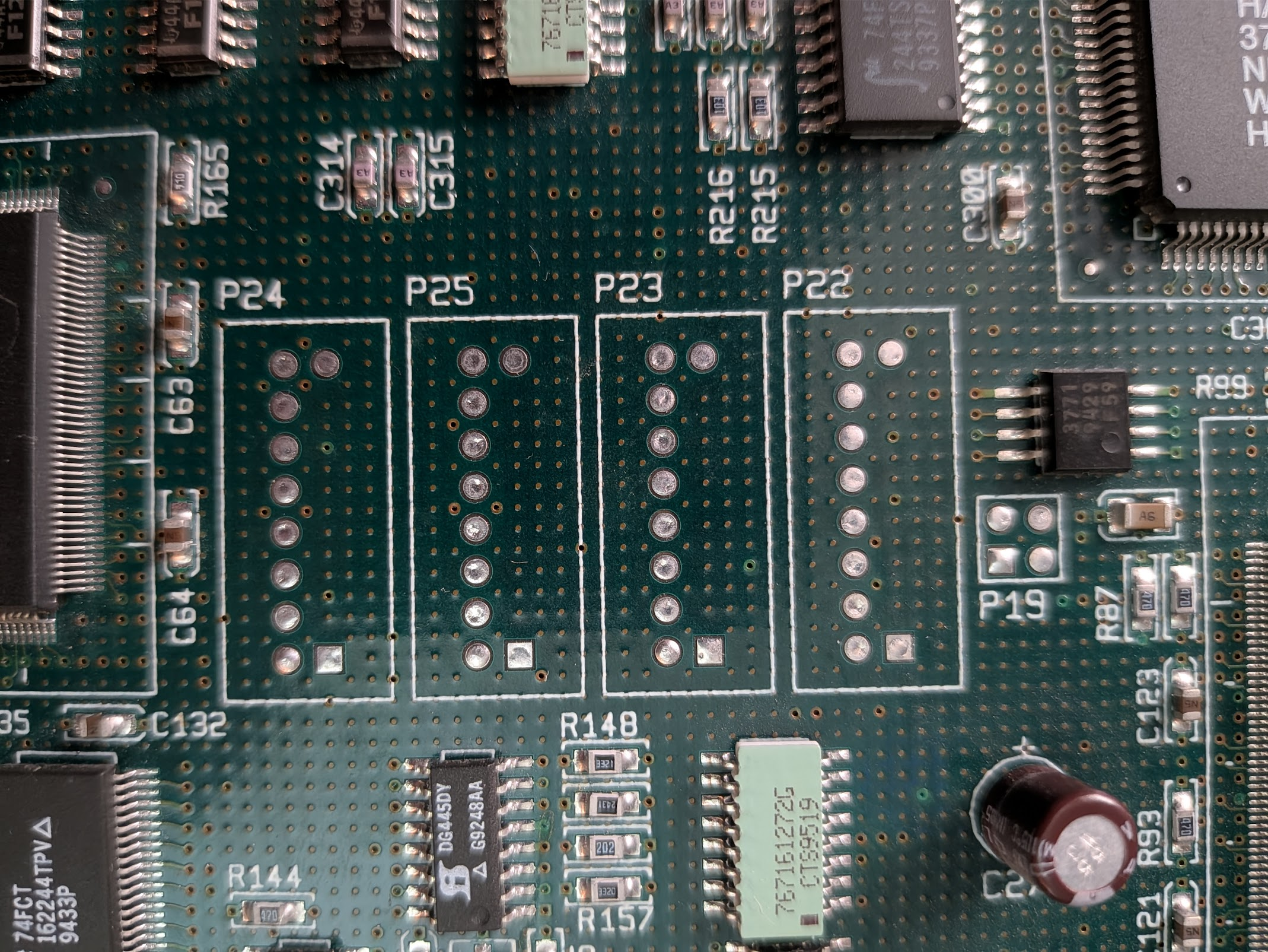 Photo of a circuit board with some unpopulated test ports. They're each 2x8 pin arrangements with one row of 8 drilled, and only the first and last pin of the second row drilled. All the drills are filled with solder, and one of the corners on the sparse row is square instead of circular. Each port has a silks border around it big enough for a 4x8 or maybe 5x10 header, suggesting a plastic housing.