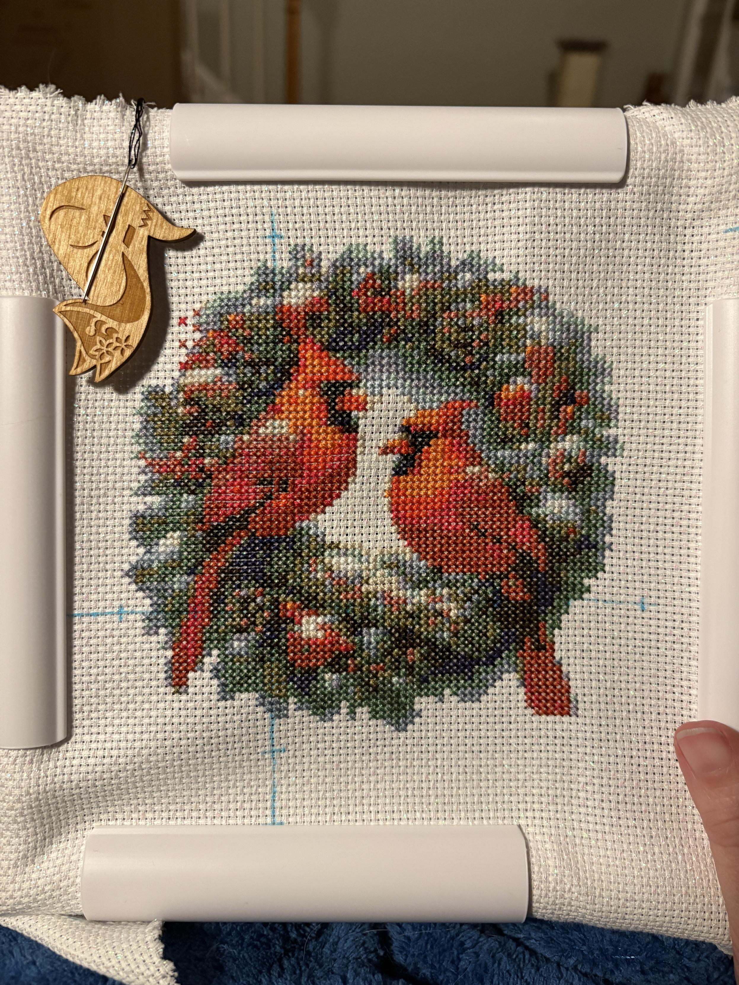 A cross stitch of two male cardinals perched on a wreath with some snow on it.