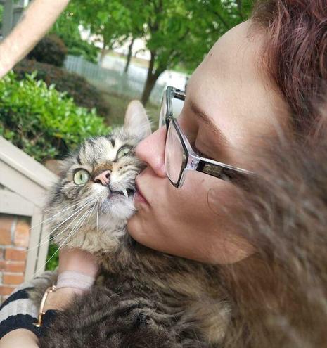 Photo of someone with pale skin, glasses, and long hair kissing a fluffy kitty they are holding outside. One of the cat's eyes is partially closed as it looks away thinking of how to escape. Kitty tried showing one fang as a deferent, but alas, they still got kissed.