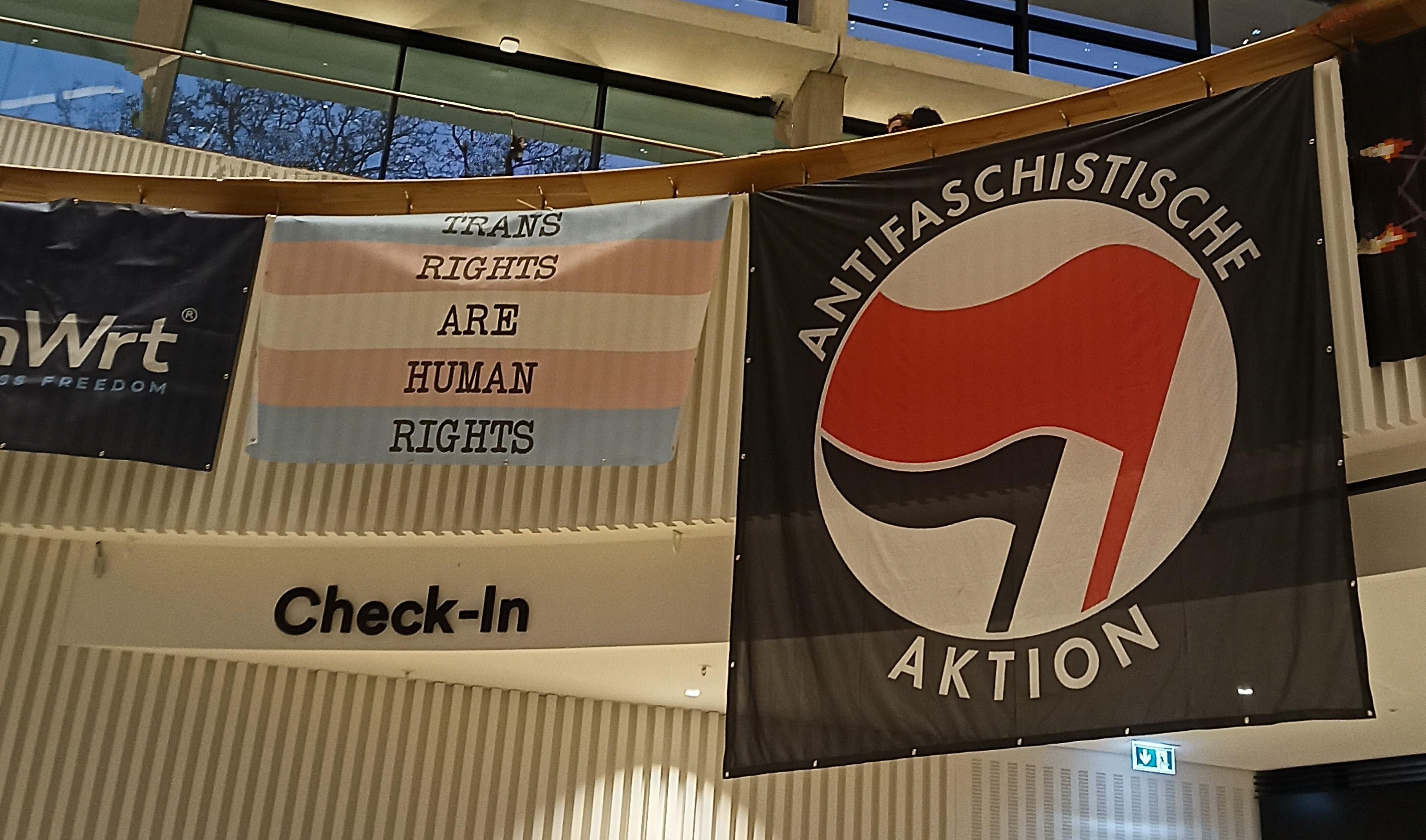 An einer Balustrade mit einem Schild Check-In hängen zwei Fahnen: eine mit hellblau und rosa mit der Aufschrift "trans rights are human rights, sowie die schwarz-rote Fahne der Antifaschistischen Aktion.