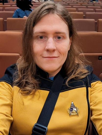​A close-up selfie of a person with shoulder-length, wavy light brown hair and a warm, confident smile. They look sharp in a crisp, mustard-yellow and black Star Trek-style uniform jacket, complete with a shiny silver Starfleet communicator badge on the chest. The background features rows of reddish-brown auditorium seats, suggesting the subject is happily attending a convention or event.
