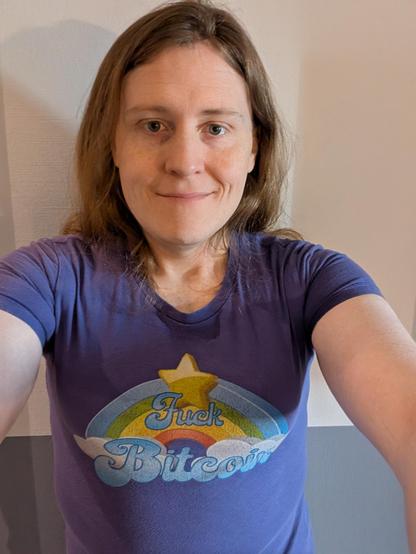 ​A warm and friendly close-up selfie of a person with long, flowing light brown hair and a gentle, kind smile. They have bright eyes and soft freckles across their cheeks, looking directly into the camera with a welcoming expression.
​The subject is wearing a purple t-shirt featuring a vibrant, retro-style graphic.

The design shows a colorful rainbow arching over soft white clouds, topped with a bright yellow star. Across the center of the rainbow, the words "Fuck Bitcoin" are written in a stylized, vintage-inspired blue and white font.