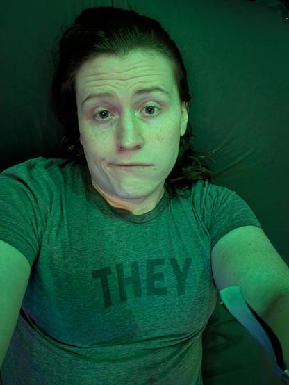 A selfie of a person with expressive eyes and light freckles, bathed in an atmospheric green light. They are looking up at the camera with a playful, wry smirk while wearing a heather grey t-shirt featuring the word THEY in dark block letters across the chest.