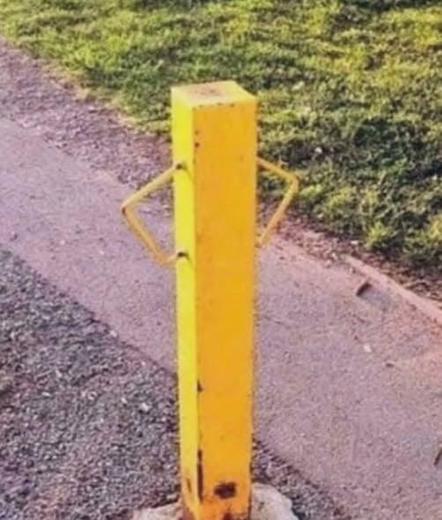 A yellow vertical bollard with two curved metal handles attached to its sides is positioned on a gravel or paved surface. The bollard shows signs of wear, including rust near its base. The surface beneath the bollard appears to be a mix of gravel and a smoother paved area, bordered by green grass in the background. The grassy area extends to the upper right of the image. There is no visible text in the image.