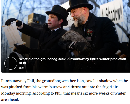 The image captures a moment from a traditional winter ceremony outdoors, likely Punxsutawney Groundhog Day. Two men in formal, old-fashioned attire dominate the scene. Both wear black top hats and long dark overcoats, along with black gloves, giving them a ceremonial, almost vintage look. The man on the left is clean-shaven and appears to be speaking or announcing something; he is holding a long sheet of paper in one hand and gesturing toward it with the other, as if presenting or reading the d…